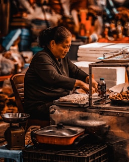 An individual sits at a food stall, preparing and arranging various dishes. The scene includes cooking utensils, trays of food items like spring rolls, and containers. The setting suggests a busy marketplace with a variety of items visible in the background.