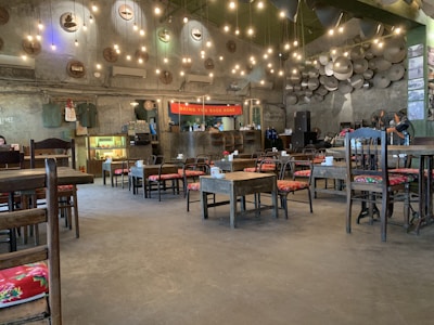 A cozy café interior with an industrial style, featuring numerous hanging lights and a high ceiling. The walls are decorated with artistic elements and clocks. The seating consists of wooden tables and chairs with colorful cushions. A counter at the back has a red banner and small shelving units.