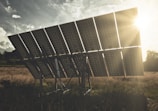 A large solar panel array stands in an open field, tilted towards the sun which is shining brightly in the background. The sky shows scattered clouds, and the sunlight creates a warm, golden glow over the landscape.