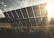 A large solar panel array stands in an open field, tilted towards the sun which is shining brightly in the background. The sky shows scattered clouds, and the sunlight creates a warm, golden glow over the landscape.