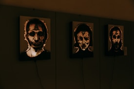 Three framed artworks are displayed side by side on a wall, each featuring dark and abstract portraits with distorted features. The lighting casts an eerie glow around the faces, enhancing their mysterious appearance.