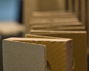 A close-up view of multiple rectangular pieces of a thick material, possibly cardboard or a composite, aligned in a row. The surfaces have a textured appearance, and the edges reveal a layered construction. The background is blurred, emphasizing the focus on the foreground elements.