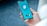 A person is holding a smartphone displaying a VPN application with a blue and teal interface. The VPN logo is centered on the screen with a checkmark and button labeled 'VPN'. The background is blurred, emphasizing the phone.