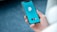 A person is holding a smartphone displaying a VPN application with a blue and teal interface. The VPN logo is centered on the screen with a checkmark and button labeled 'VPN'. The background is blurred, emphasizing the phone.