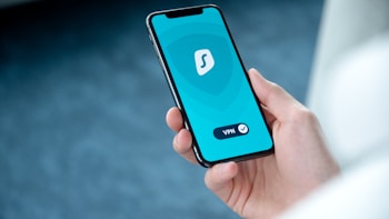 A person is holding a smartphone displaying a VPN application with a blue and teal interface. The VPN logo is centered on the screen with a checkmark and button labeled 'VPN'. The background is blurred, emphasizing the phone.