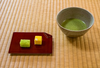 A ceramic bowl containing green matcha tea is placed on a tatami mat. Beside it, a red rectangular plate holds two small sponge cakes, one green and one yellow. The setting has a serene and traditional aesthetic.