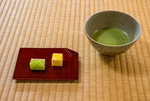 A ceramic bowl containing green matcha tea is placed on a tatami mat. Beside it, a red rectangular plate holds two small sponge cakes, one green and one yellow. The setting has a serene and traditional aesthetic.