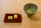 A ceramic bowl containing green matcha tea is placed on a tatami mat. Beside it, a red rectangular plate holds two small sponge cakes, one green and one yellow. The setting has a serene and traditional aesthetic.