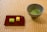 A ceramic bowl containing green matcha tea is placed on a tatami mat. Beside it, a red rectangular plate holds two small sponge cakes, one green and one yellow. The setting has a serene and traditional aesthetic.