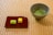 A ceramic bowl containing green matcha tea is placed on a tatami mat. Beside it, a red rectangular plate holds two small sponge cakes, one green and one yellow. The setting has a serene and traditional aesthetic.