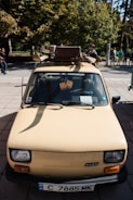 A vintage compact car with a cream color is parked outdoors. It has a roof rack holding a suitcase and some luggage. The view is from the front, showing the car's details like headlights and license plate. Hanging from the rear-view mirror are two fuzzy dice. People are visible in the background along with trees and a park setting.