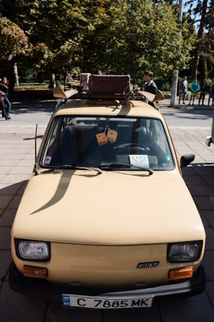 A vintage compact car with a cream color is parked outdoors. It has a roof rack holding a suitcase and some luggage. The view is from the front, showing the car's details like headlights and license plate. Hanging from the rear-view mirror are two fuzzy dice. People are visible in the background along with trees and a park setting.