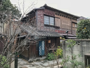 A traditional wooden Japanese house with a tiled roof surrounded by trees and a small garden area. The exterior features sliding windows and wooden slats, contributing to a rustic aesthetic. A blue noren (fabric divider) hangs near the entrance, and a small citrus tree is visible to the right.