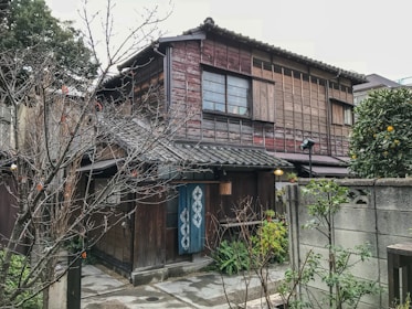 A traditional wooden Japanese house with a tiled roof surrounded by trees and a small garden area. The exterior features sliding windows and wooden slats, contributing to a rustic aesthetic. A blue noren (fabric divider) hangs near the entrance, and a small citrus tree is visible to the right.