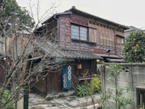 A traditional wooden Japanese house with a tiled roof surrounded by trees and a small garden area. The exterior features sliding windows and wooden slats, contributing to a rustic aesthetic. A blue noren (fabric divider) hangs near the entrance, and a small citrus tree is visible to the right.