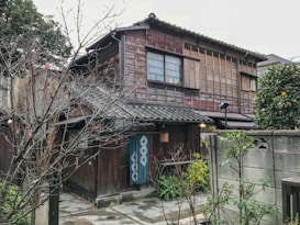 A traditional wooden Japanese house with a tiled roof surrounded by trees and a small garden area. The exterior features sliding windows and wooden slats, contributing to a rustic aesthetic. A blue noren (fabric divider) hangs near the entrance, and a small citrus tree is visible to the right.