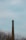 A tall, narrow brick chimney rises above leafless trees under a pale, overcast sky. The structure has metal ladders affixed to its side, extending upward toward the top.