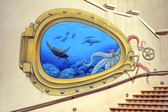 A mural painted on a building wall depicting an underwater scene as seen through a large brass porthole. Within the porthole are several marine animals including a sea turtle, a school of fish, dolphins, and an octopus with its tentacles extending outside the frame. The mural features vibrant coral in the background and is bordered by metallic hinges and rivets.