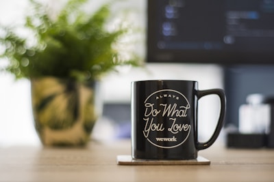 A black mug with white text saying 'Always Do What You Love - wework' is placed on a wooden table. A blurred potted plant and a monitor are visible in the background, adding a cozy and professional atmosphere.