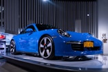 A sleek blue sports car is displayed on a platform under bright lights in a modern showroom. The car features a New York license plate that reads 'PCA 60TH' and is surrounded by other promotional images and displays.