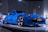 A sleek blue sports car is displayed on a platform under bright lights in a modern showroom. The car features a New York license plate that reads 'PCA 60TH' and is surrounded by other promotional images and displays.