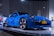 A sleek blue sports car is displayed on a platform under bright lights in a modern showroom. The car features a New York license plate that reads 'PCA 60TH' and is surrounded by other promotional images and displays.
