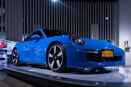 A sleek blue sports car is displayed on a platform under bright lights in a modern showroom. The car features a New York license plate that reads 'PCA 60TH' and is surrounded by other promotional images and displays.