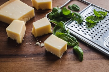 Several cubes of a light yellow cheese are placed on a wooden surface alongside scattered fresh green basil leaves. A metal cheese grater lies nearby.