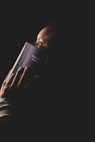 A person holding a book with an intricate cover and gold text close to their face. The lighting focuses on the person's profile and the book, creating a dramatic and intimate atmosphere against a dark background.