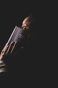 A person holding a book with an intricate cover and gold text close to their face. The lighting focuses on the person's profile and the book, creating a dramatic and intimate atmosphere against a dark background.