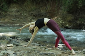 A person is performing a yoga pose in an outdoor setting by a flowing river. The individual is in a graceful backbend with one arm extended toward the ground. They are dressed in a black top and burgundy leggings. The background includes greenery and the gentle movement of water over rocks.