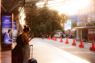 A person with a backpack stands on a platform glancing at a smartphone while their luggage is beside them. The scene is lit by a soft, warm light that creates long shadows. Orange traffic cones line the edge of the walkway, and several informational advertisements are visible in the background. The surrounding area includes greenery and a few parked vehicles.
