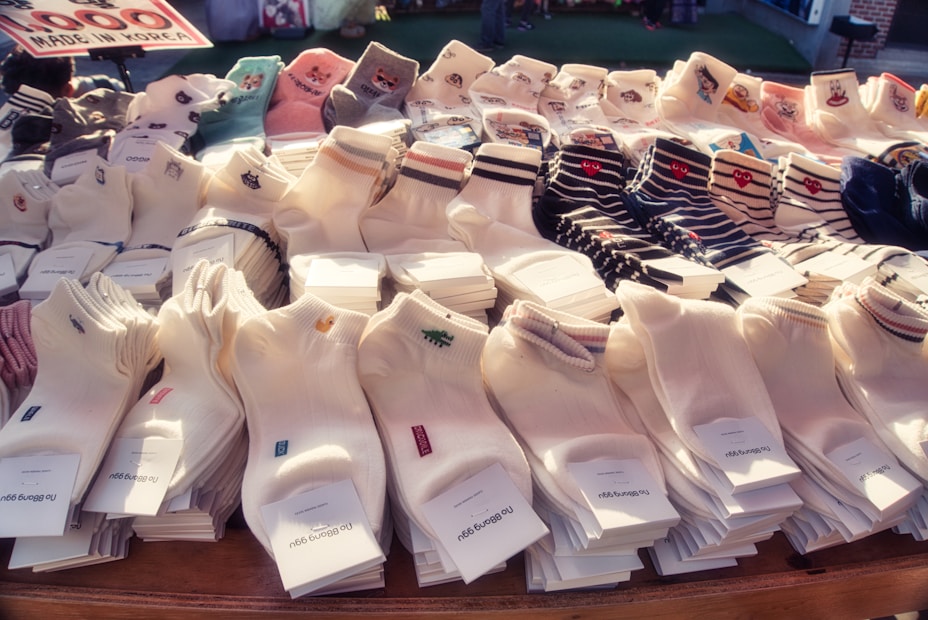A display of various socks neatly arranged in rows. The socks come in different patterns and colors, including solid white, striped, and those with small embroidered designs. A sign indicates they are made in Korea, and the price is noted as 1000. The setting appears to be a market or a store with a variety of socks on offer.