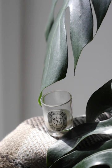 A close-up of a softly glowing Vespera Bloom candle in minimalist deep aubergine glass, casting gentle shadows on a crisp white surface.