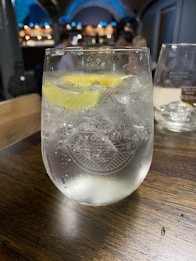 A clear glass filled with a fizzy drink and ice cubes, garnished with a slice of lemon. It is placed on a wooden table, with a blurred background showing other glasses and a softly lit bar area.