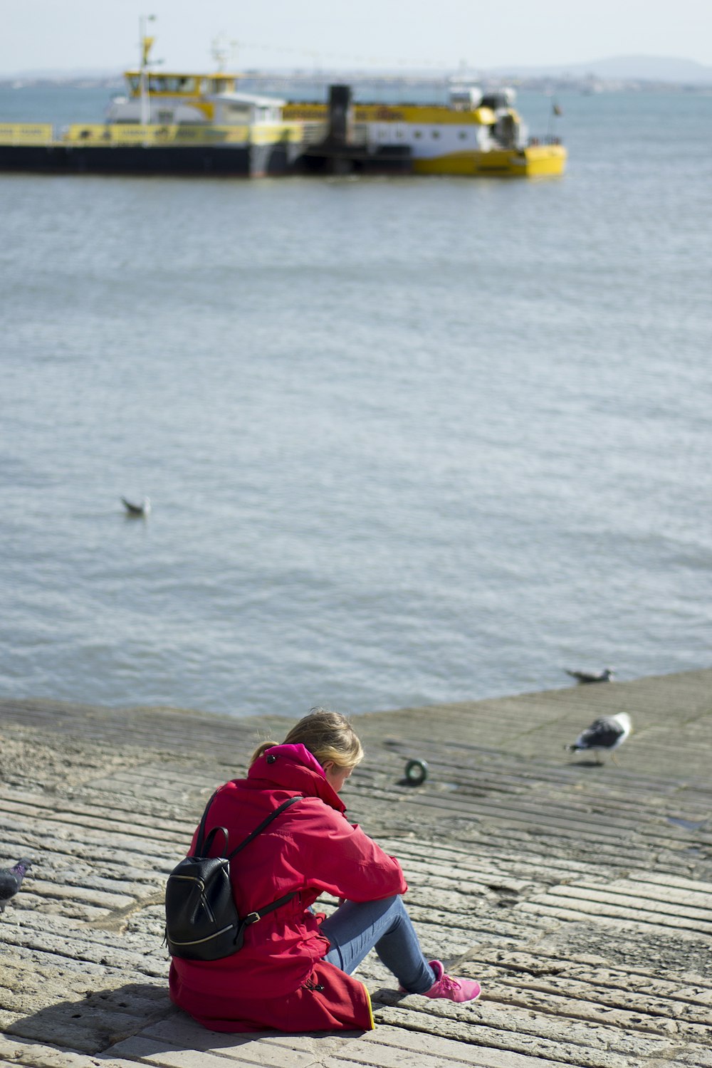 woman in red hoodie sitting on brown wooden dock looking at sea during daytime