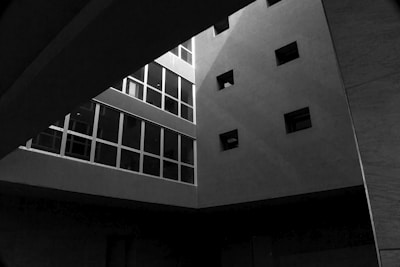 An architectural scene with a modern building interior featuring geometric shapes and strong contrasts of light and shadow. Several rectangular windows are visible, creating a grid-like pattern on one wall. The structure gives a feeling of depth and minimalism due to the interplay of light and architectural elements.