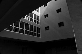 An architectural scene with a modern building interior featuring geometric shapes and strong contrasts of light and shadow. Several rectangular windows are visible, creating a grid-like pattern on one wall. The structure gives a feeling of depth and minimalism due to the interplay of light and architectural elements.