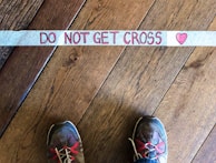 A pair of shoes standing on wooden floorboards next to a strip of tape with the words 'DO NOT GET CROSS' written in red letters. A heart symbol is drawn next to the text.