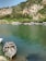 A serene river landscape featuring a small, wooden rowing boat tied at the water's edge. Across the green river, several tour boats are docked beside a lush, tree-covered shoreline. Beyond the trees, ancient rock tombs are carved into the hillside, framed by rugged cliffs and dense greenery.
