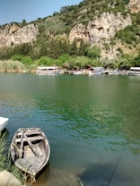 A serene river landscape featuring a small, wooden rowing boat tied at the water's edge. Across the green river, several tour boats are docked beside a lush, tree-covered shoreline. Beyond the trees, ancient rock tombs are carved into the hillside, framed by rugged cliffs and dense greenery.