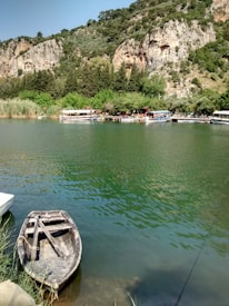 A serene river landscape featuring a small, wooden rowing boat tied at the water's edge. Across the green river, several tour boats are docked beside a lush, tree-covered shoreline. Beyond the trees, ancient rock tombs are carved into the hillside, framed by rugged cliffs and dense greenery.