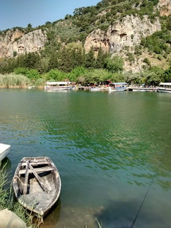 A serene river landscape featuring a small, wooden rowing boat tied at the water's edge. Across the green river, several tour boats are docked beside a lush, tree-covered shoreline. Beyond the trees, ancient rock tombs are carved into the hillside, framed by rugged cliffs and dense greenery.