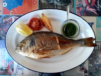 A whole grilled fish is served on a white oval plate with a grilled tomato, a wedge of lemon, a piece of roasted onion, and a small bowl of green sauce with a spoon on the side. The plate rests on a colorful table covered with graphic designs and texts.