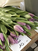 A bundle of light purple tulips with green leaves is laid out on a wooden surface. There is a handwritten sign nearby indicating a price of 10 with an unclear unit or currency.