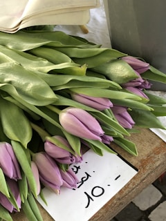 A bundle of light purple tulips with green leaves is laid out on a wooden surface. There is a handwritten sign nearby indicating a price of 10 with an unclear unit or currency.