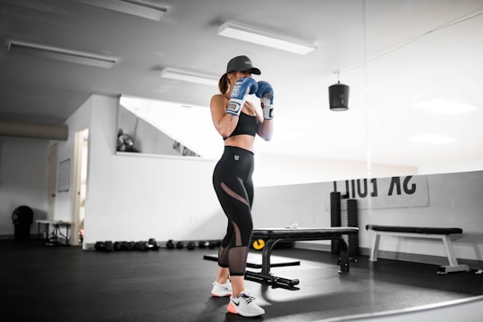 A person wearing athletic wear and a cap is in a gym setting, practicing boxing with blue gloves. The gym is well-lit and features fitness equipment such as dumbbells and benches. The person appears focused and is positioned in front of a mirrored wall.