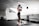 A person wearing athletic wear and a cap is in a gym setting, practicing boxing with blue gloves. The gym is well-lit and features fitness equipment such as dumbbells and benches. The person appears focused and is positioned in front of a mirrored wall.