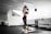 A person wearing athletic wear and a cap is in a gym setting, practicing boxing with blue gloves. The gym is well-lit and features fitness equipment such as dumbbells and benches. The person appears focused and is positioned in front of a mirrored wall.