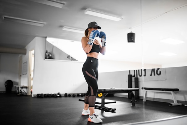 A person wearing athletic wear and a cap is in a gym setting, practicing boxing with blue gloves. The gym is well-lit and features fitness equipment such as dumbbells and benches. The person appears focused and is positioned in front of a mirrored wall.